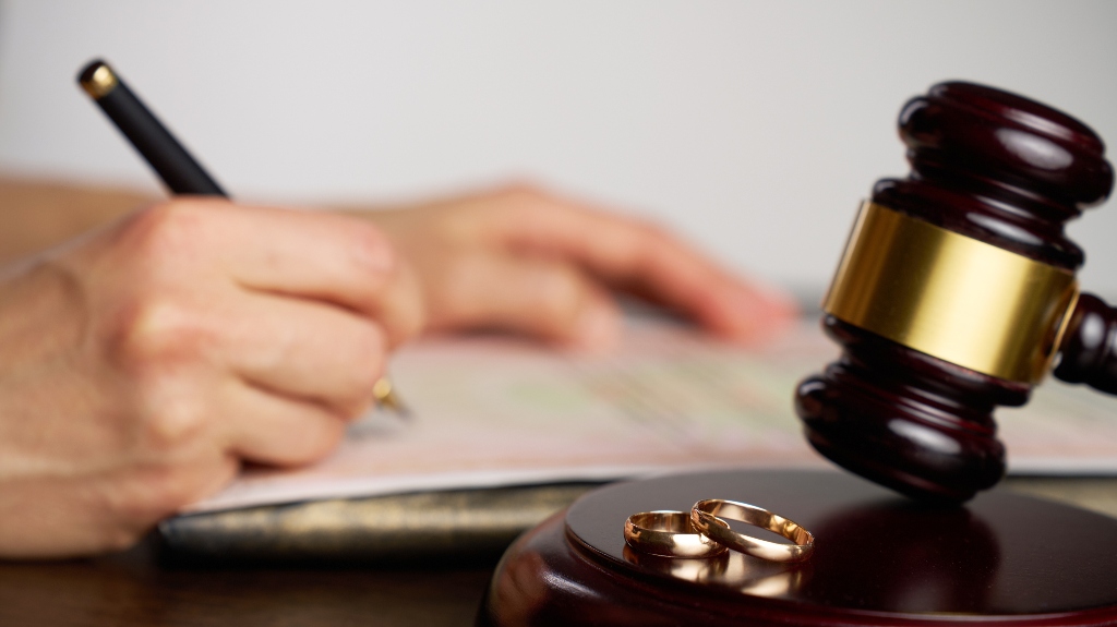 person signing divorce document, with a judge gavel and wedding rings in focus