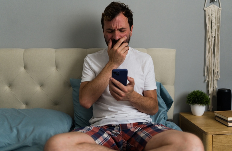 Upset man sitting on bed reading bad news on smartphone and crying